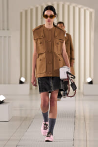 Model walks a fashion runway in a tan utility vest with large pockets, a black leather mini skirt, pink sneakers, gray socks, round sunglasses, and a white handbag with a red chain accessory.