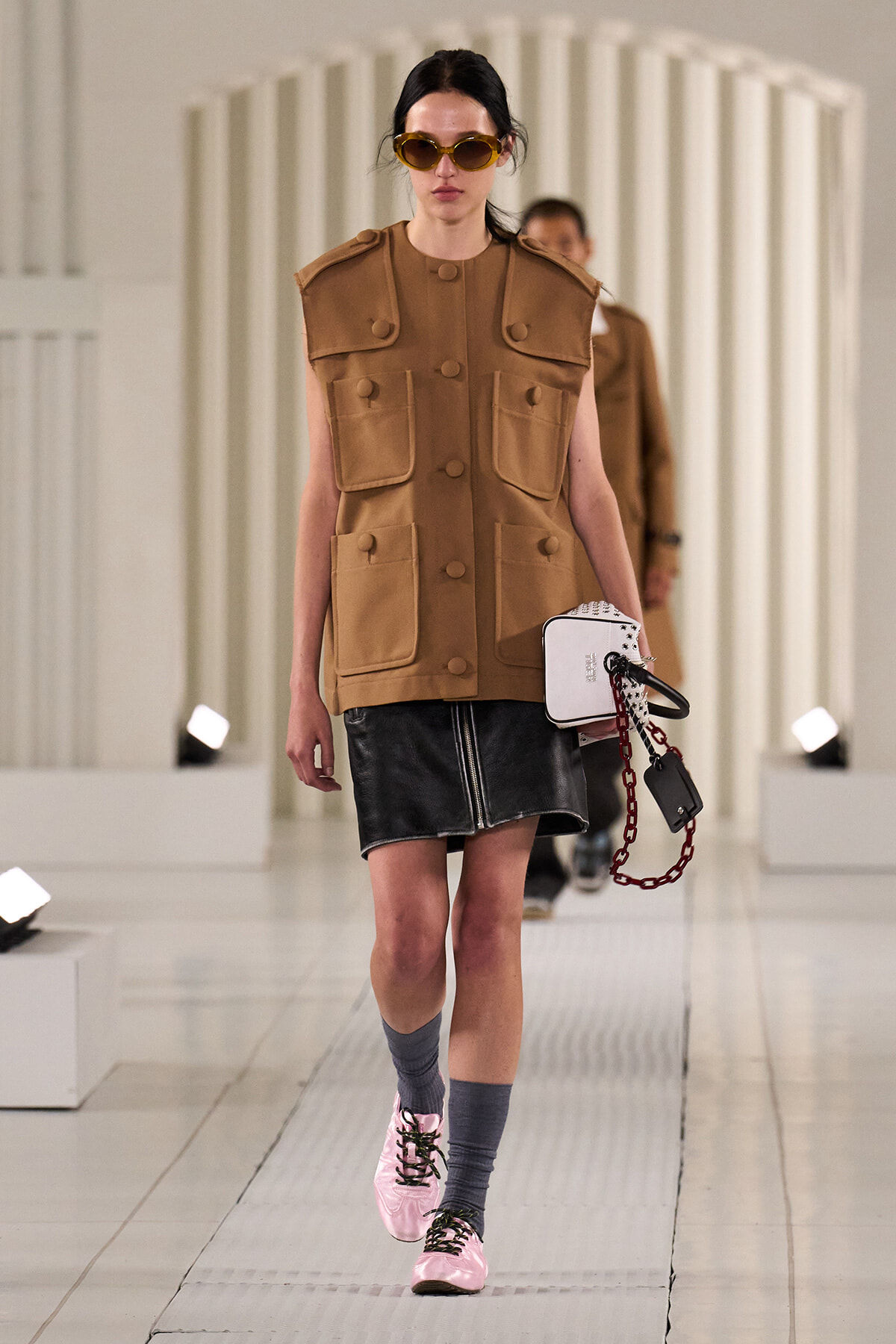 Model walks a fashion runway in a tan utility vest with large pockets, a black leather mini skirt, pink sneakers, gray socks, round sunglasses, and a white handbag with a red chain accessory.