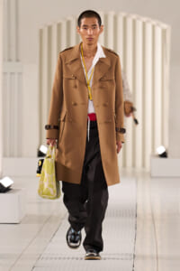 Male model walks runway in a camel trench coat over a white shirt and black trousers, carrying a light-green patterned tote bag.