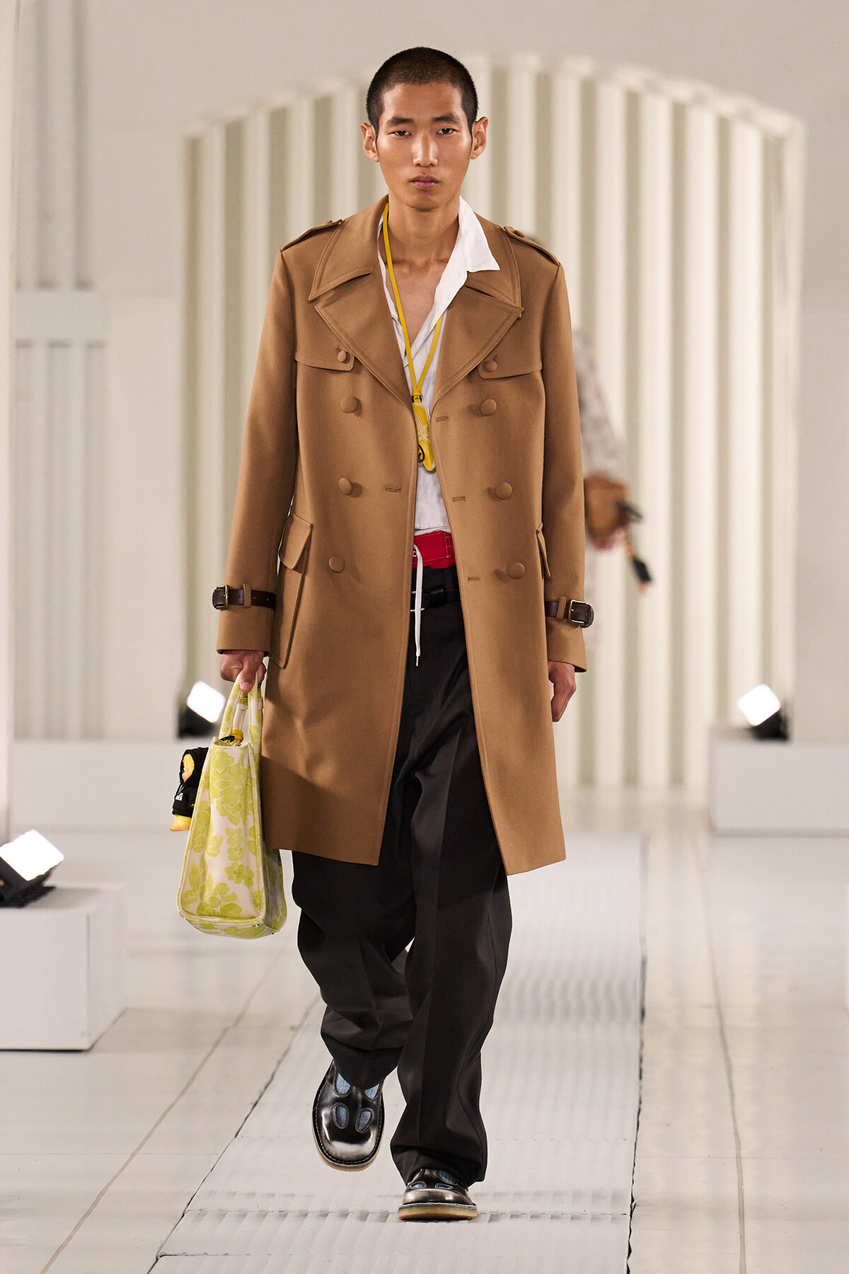 Male model walks runway in a camel trench coat over a white shirt and black trousers, carrying a light-green patterned tote bag.
