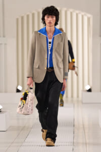 Male model walking a runway in a beige blazer over a blue zip jacket, black pants, and tan shoes, carrying a printed tote bag with flowers
