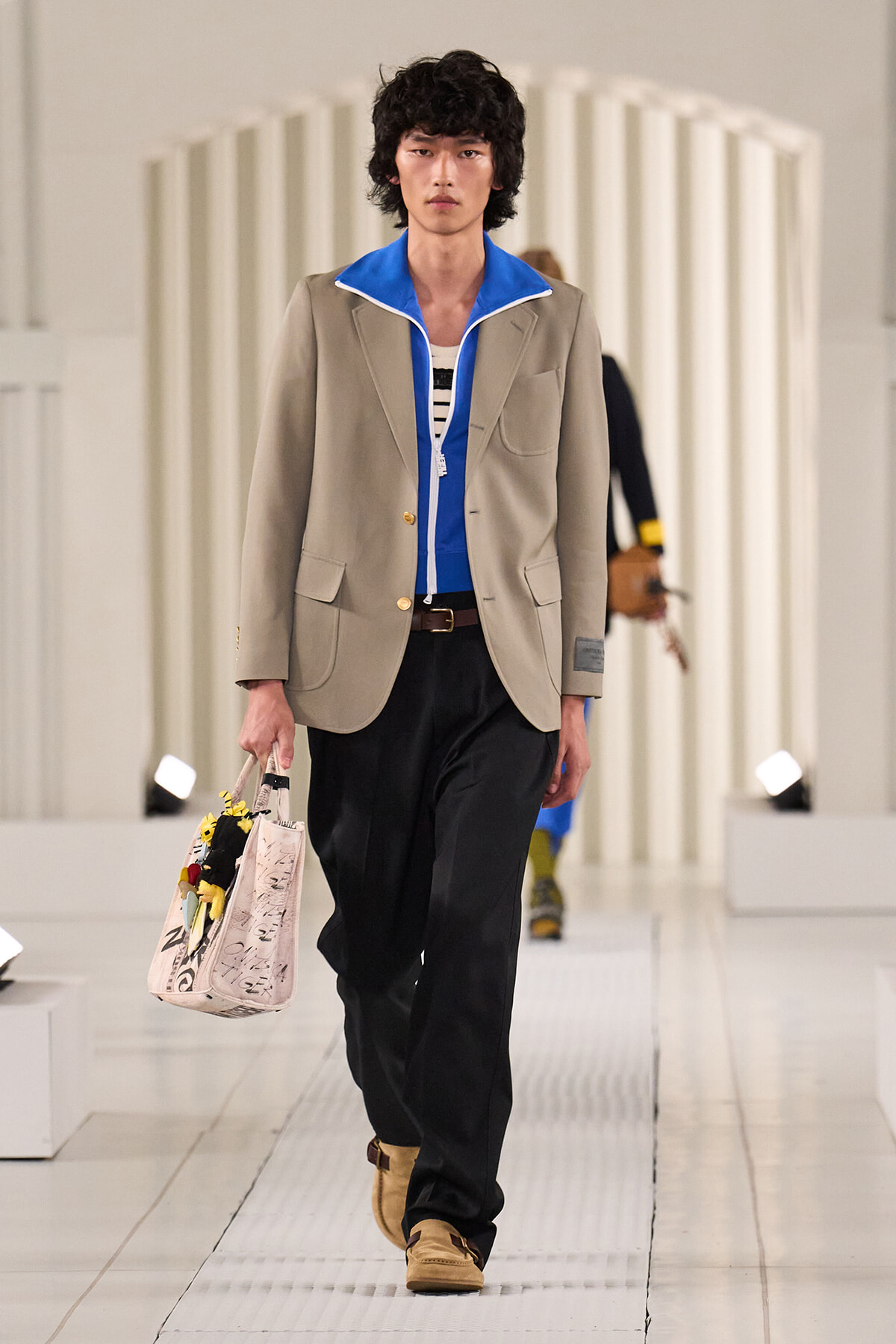 Male model walking a runway in a beige blazer over a blue zip jacket, black pants, and tan shoes, carrying a printed tote bag with flowers
