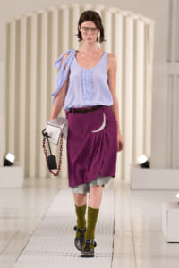 Model walking runway in a light blue sleeveless top with a shoulder tie, rust purple draped skirt over gray shorts, olive-green socks, black sandals, and a white bag with a red chain.