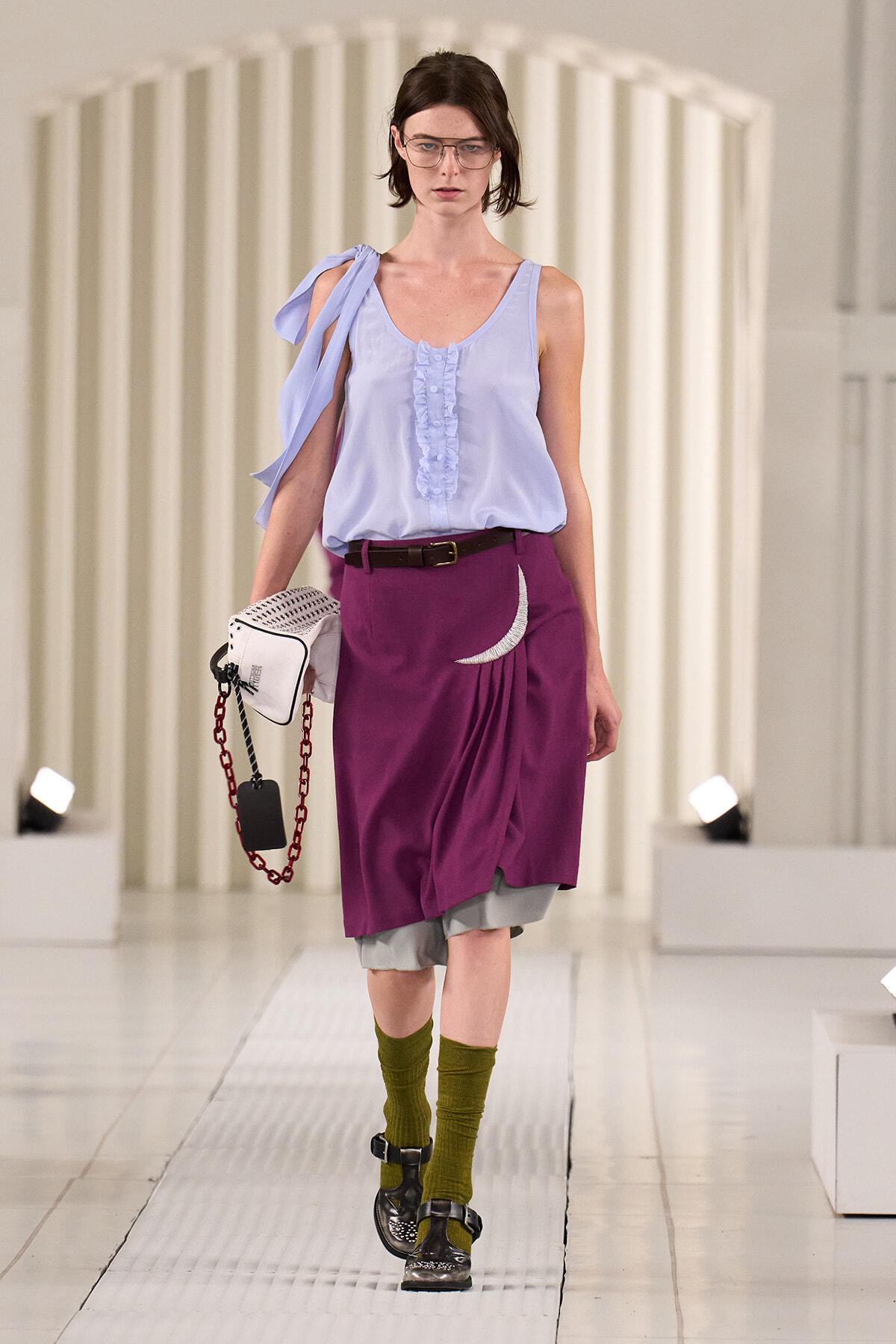 Model walking runway in a light blue sleeveless top with a shoulder tie, rust purple draped skirt over gray shorts, olive-green socks, black sandals, and a white bag with a red chain.