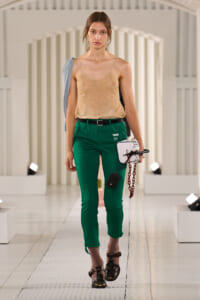 Female model walking a fashion runway in a beige lace camisole and green tailored pants, carrying a white structured bag with a red chain and wearing brown sandals.
