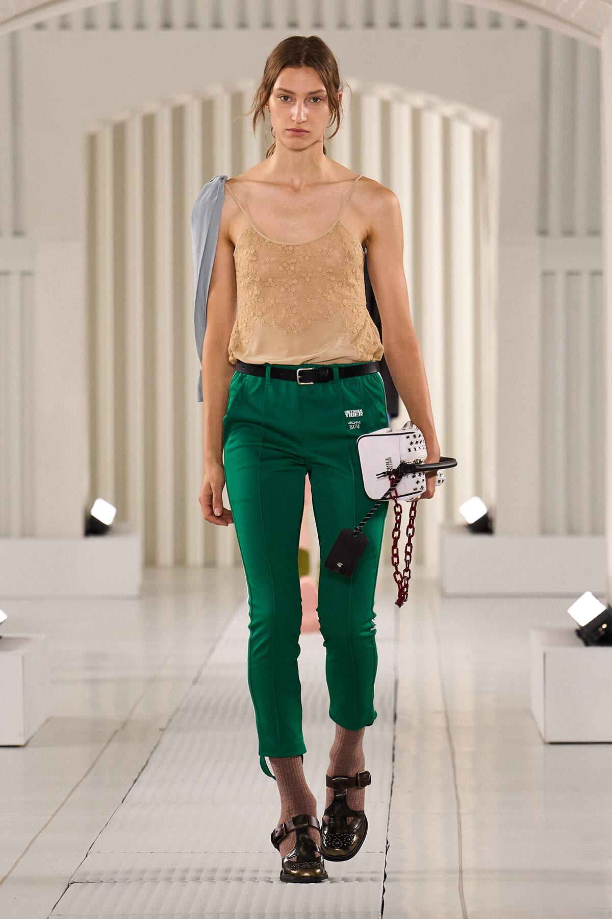 Female model walking a fashion runway in a beige lace camisole and green tailored pants, carrying a white structured bag with a red chain and wearing brown sandals.