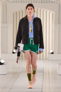 Model walking a runway in a light blue shirt, black zip jacket, green shorts, pink sneakers, and olive socks, with a yellow badge on a lanyard at the neck.
