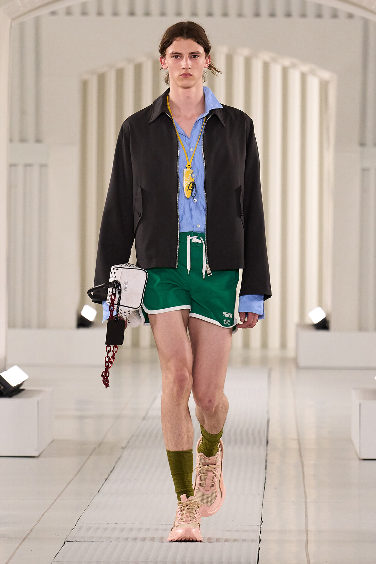 Model walking a runway in a light blue shirt, black zip jacket, green shorts, pink sneakers, and olive socks, with a yellow badge on a lanyard at the neck.