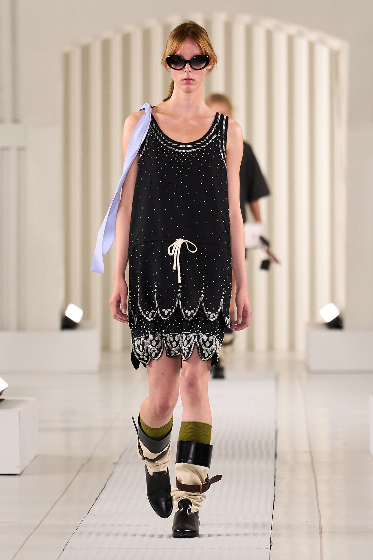 Model on a runway wearing a black sleeveless dotted dress with white embroidery at the hem, sunglasses, and slouchy boots.