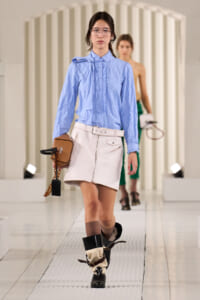 Model walking a fashion runway in a light blue ruffled blouse and white mini skirt, carrying a tan mini bag with a chain accessory.