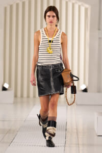 Model walking a runway in a white and navy striped sleeveless top with a black leather mini skirt, tan bag with chain, and stacked boots with leg wraps.