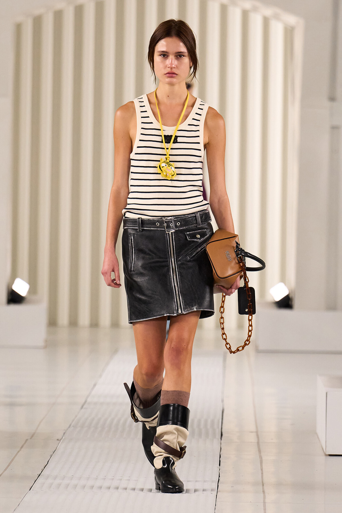 Model walking a runway in a white and navy striped sleeveless top with a black leather mini skirt, tan bag with chain, and stacked boots with leg wraps.