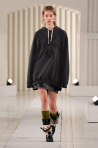 Model walking a fashion runway in an oversized black hoodie with white drawstrings, a black leather mini skirt, olive socks, and heeled black boots.