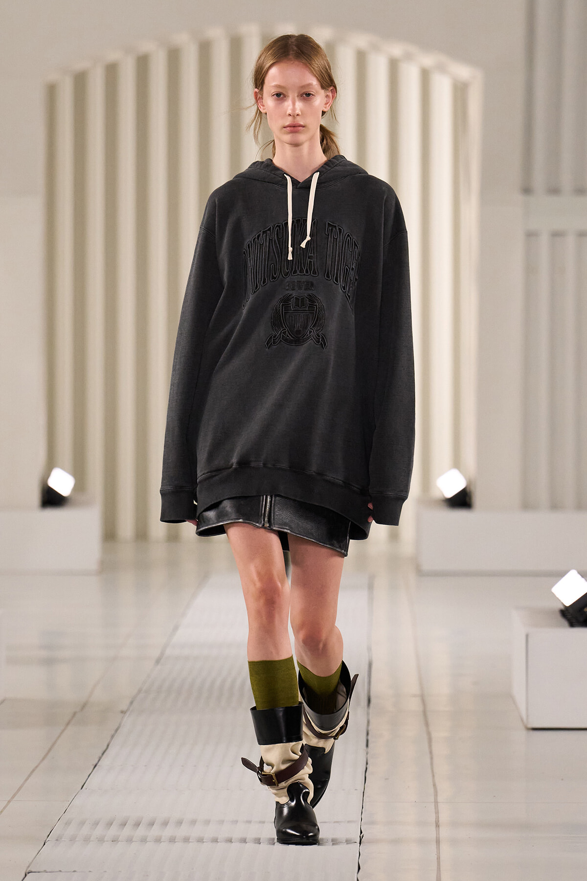 Model walking a fashion runway in an oversized black hoodie with white drawstrings, a black leather mini skirt, olive socks, and heeled black boots.