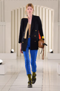 Model walking a runway in a black double-breasted blazer with gold buttons, tan scarf, electric blue pants, olive socks, chunky black shoes, and a brown bag with a chain.