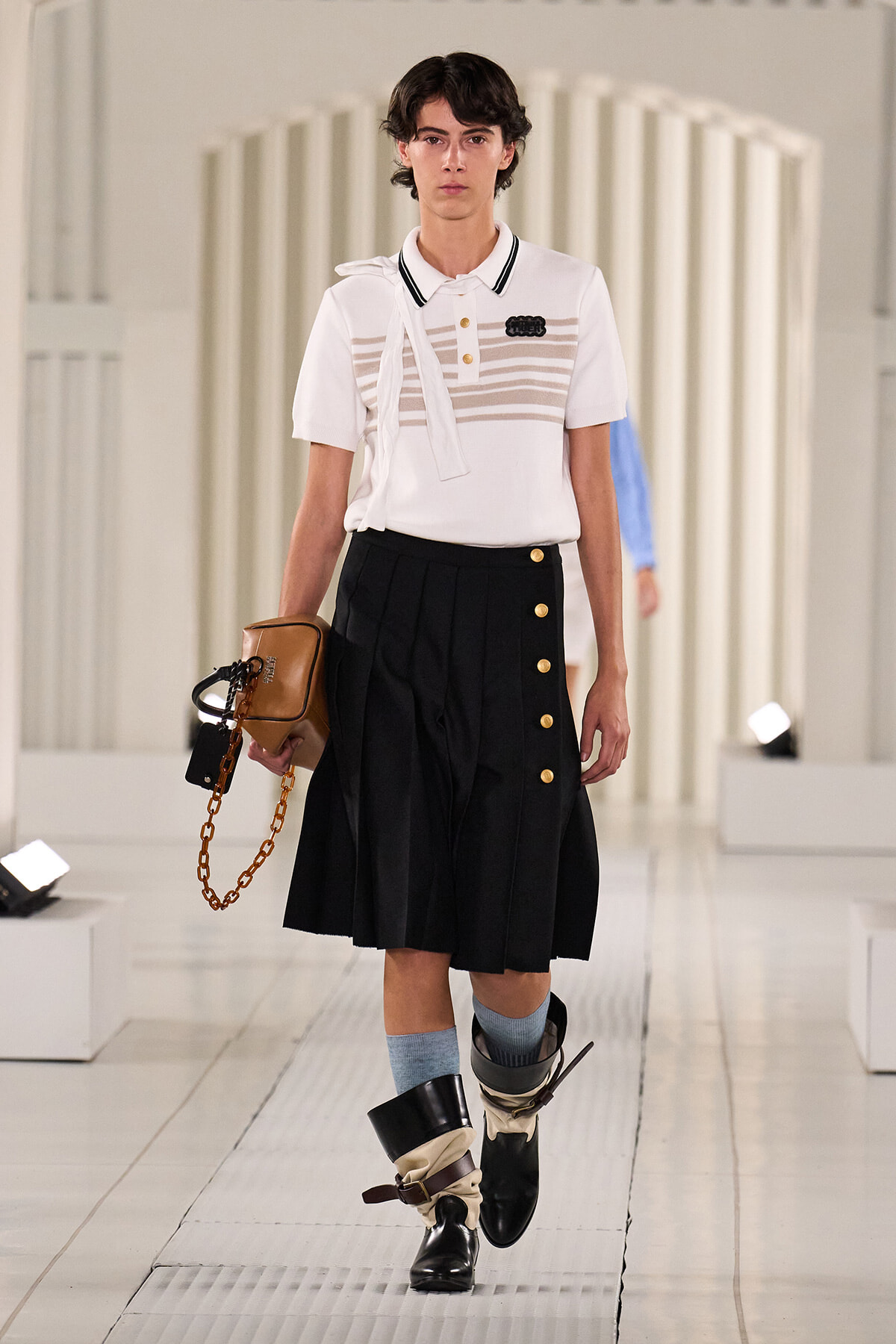 Model walking a fashion runway in a white polo with striped chest panel, black pleated skirt, and knee-high boots with layered wraps; brown handbag with gold chain accessory