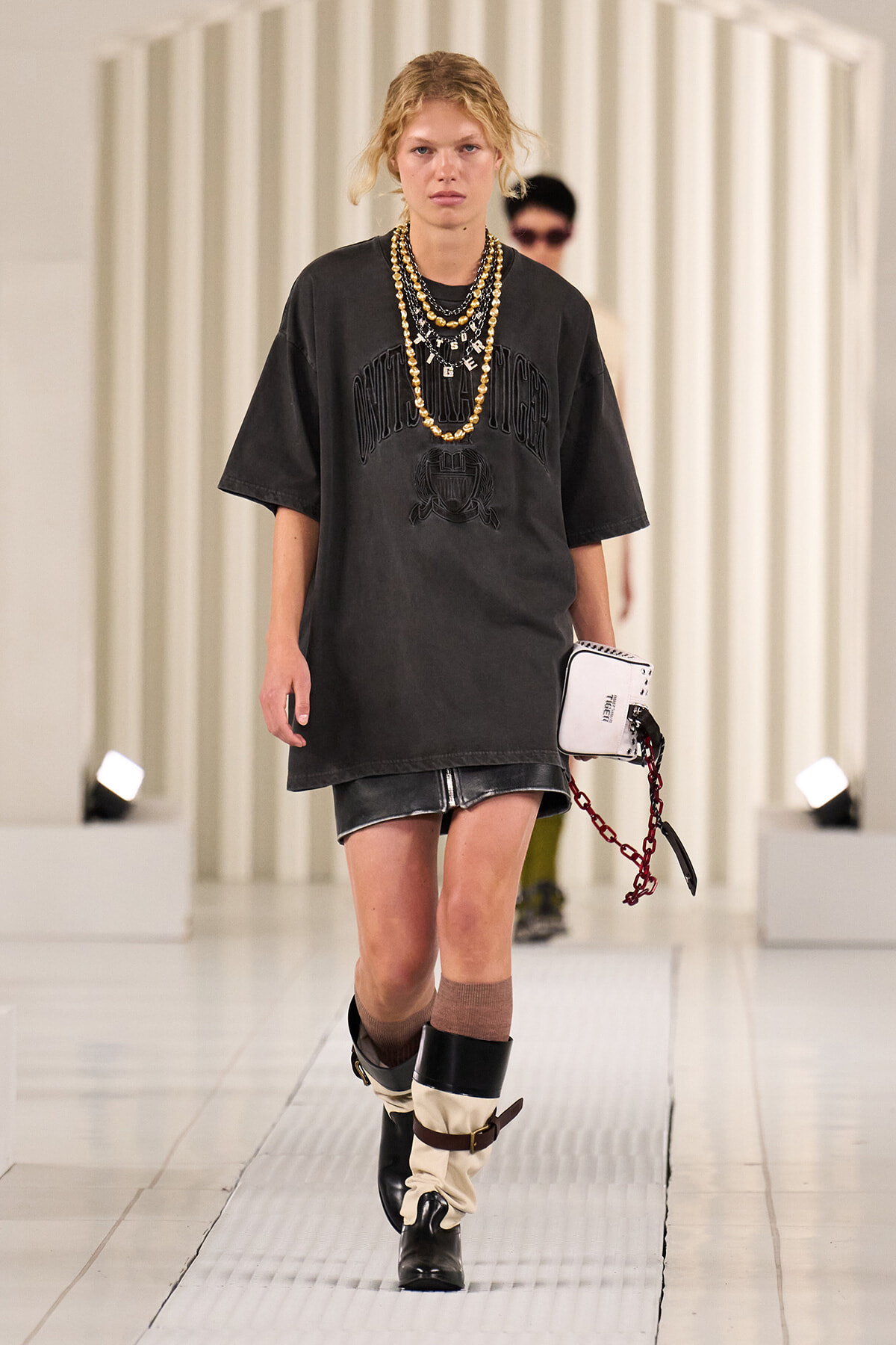 Model walking the runway in an oversized black graphic tee, shorts, layered gold necklaces, and two-tone slouch boots, carrying a white clutch with a red chain accessory