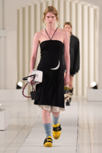 Model on a runway wearing a black strapless dress, pale underlayer, blue socks, and bright yellow sandals, carrying a white handbag with a red chain.