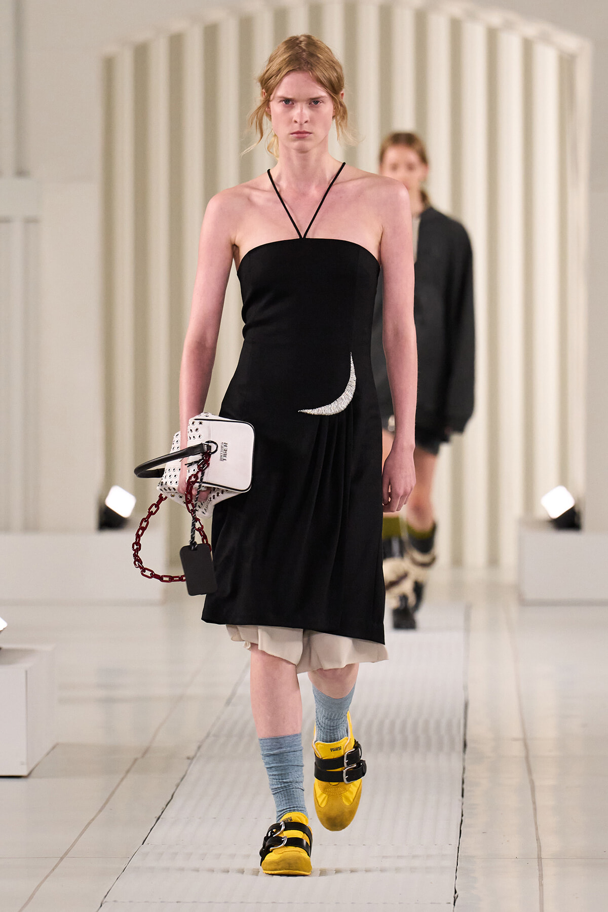 Model on a runway wearing a black strapless dress, pale underlayer, blue socks, and bright yellow sandals, carrying a white handbag with a red chain.