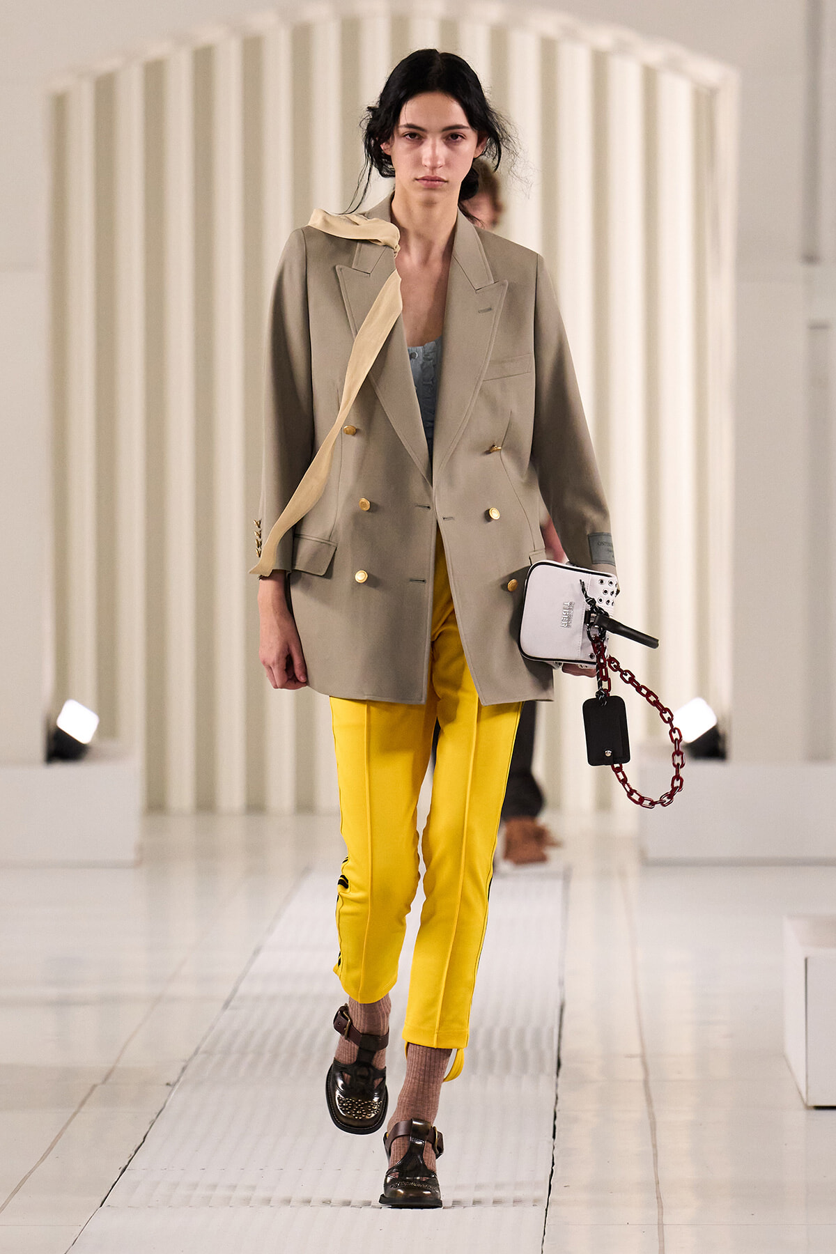 Model walks the runway in a beige double-breasted blazer, blue top, and bright yellow trousers, carrying a white purse with a chain accessory.