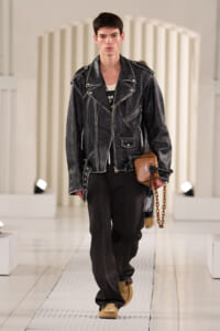 Male model on a runway wearing a black distressed leather biker jacket, black pants, tan boots, and a small tan pouch with a chain.