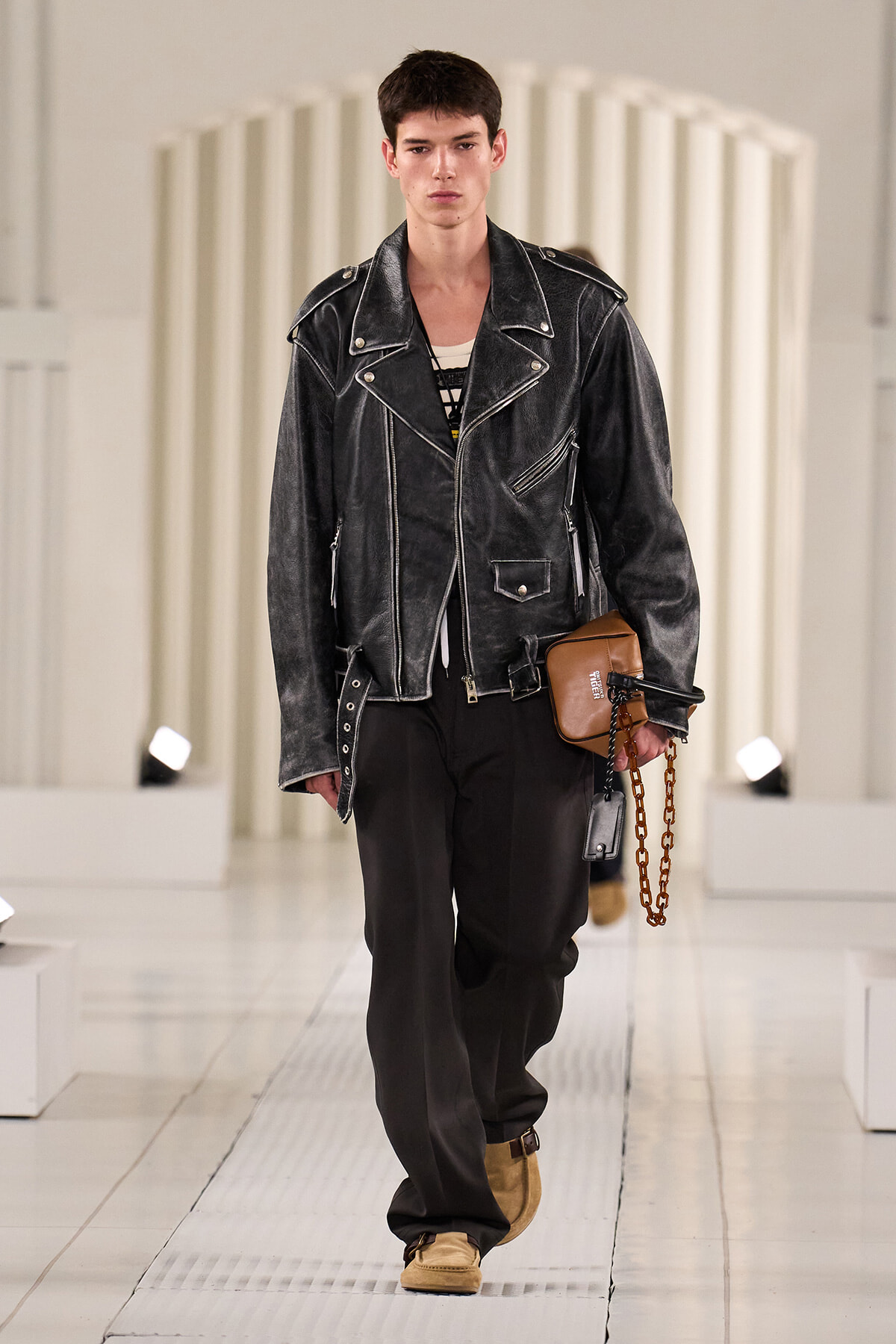 Male model on a runway wearing a black distressed leather biker jacket, black pants, tan boots, and a small tan pouch with a chain.