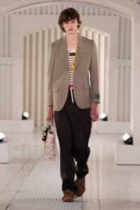 Male model walks a runway in a beige blazer over a striped shirt, black pants, brown fringe loafers, yellow lanyard, and carries a light tote bag with a small plush accessory.