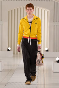 Male model walking a fashion runway in a bright yellow zip-up jacket with black pants and a beige tote bag.