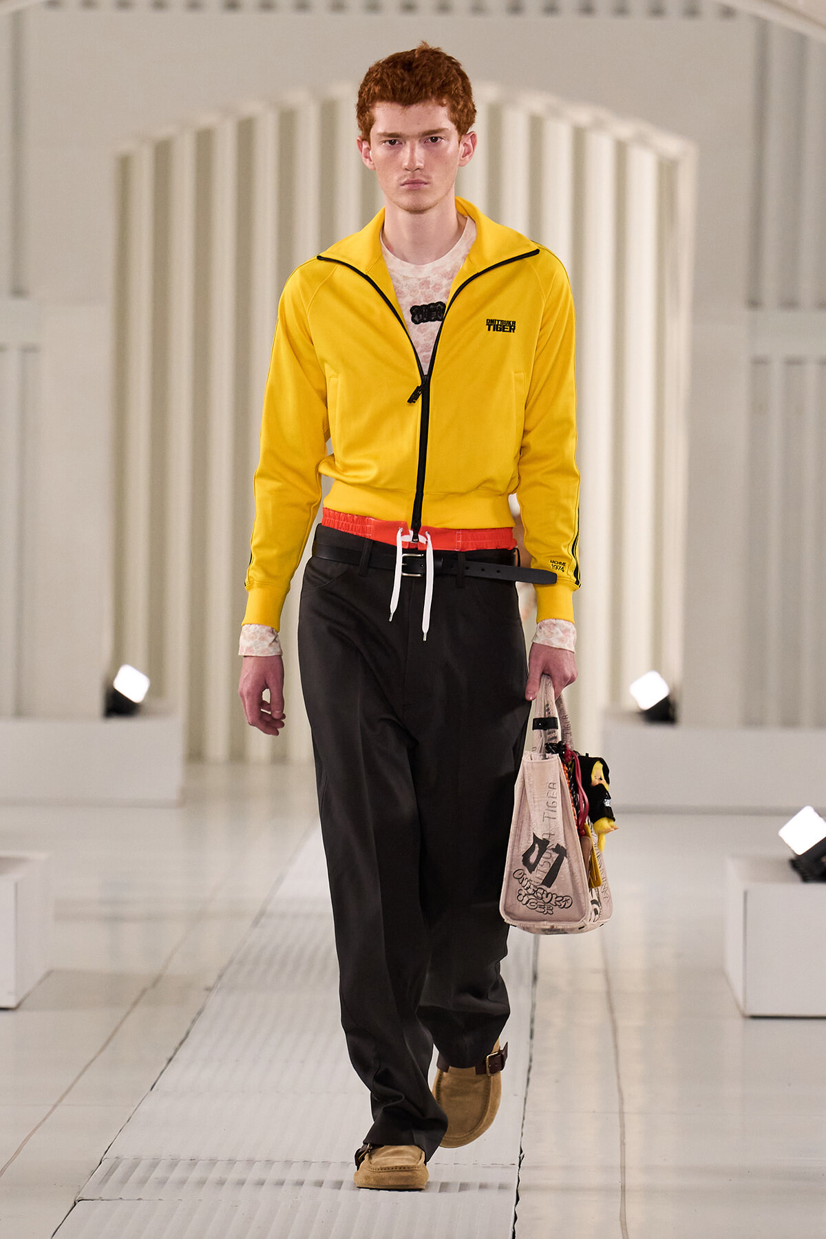 Male model walking a fashion runway in a bright yellow zip-up jacket with black pants and a beige tote bag.