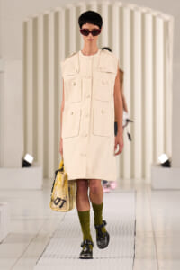 Model on a white runway wearing a cream sleeveless utility coat with large front pockets and oversized buttons, carrying a yellow graphic tote.