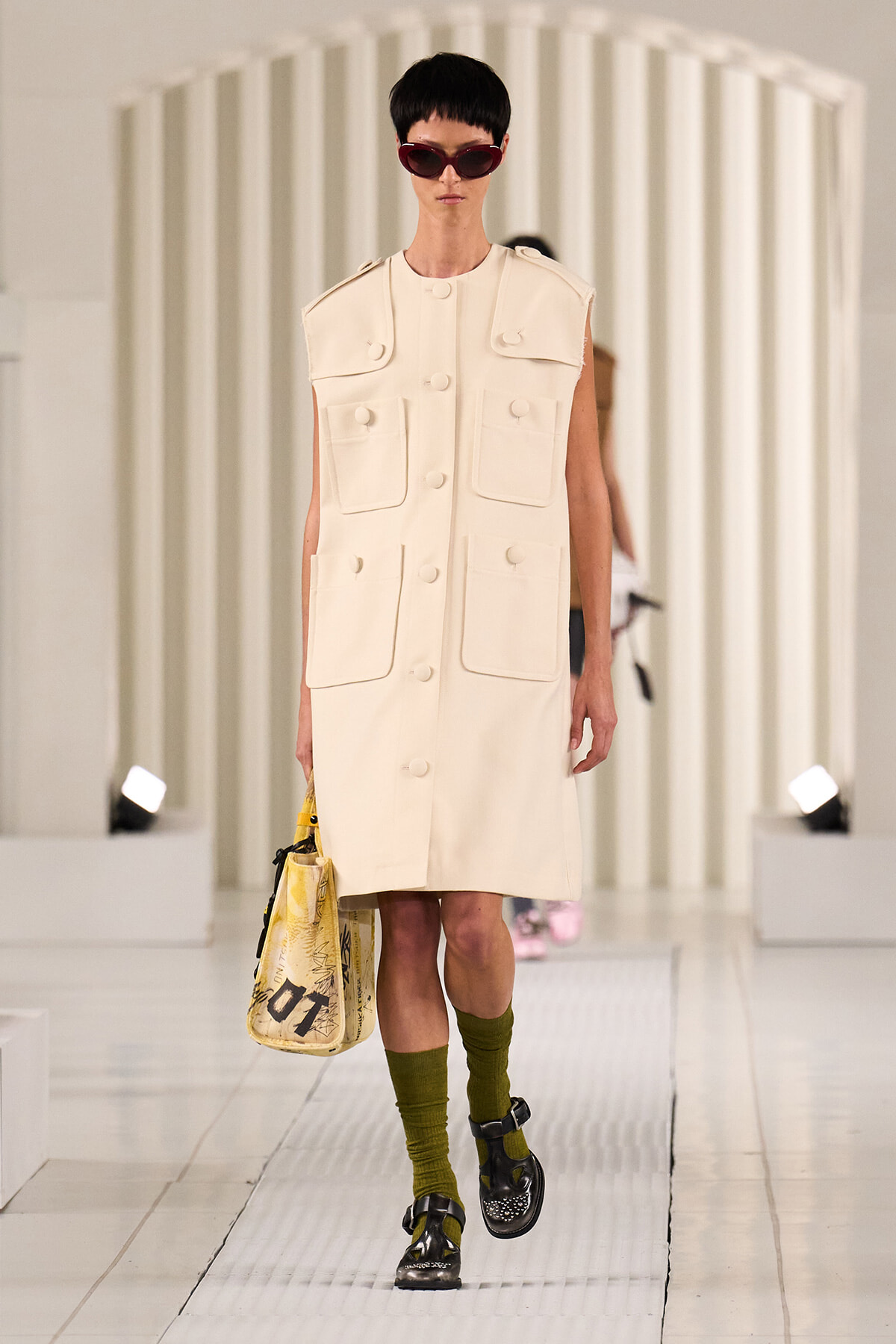 Model on a white runway wearing a cream sleeveless utility coat with large front pockets and oversized buttons, carrying a yellow graphic tote.