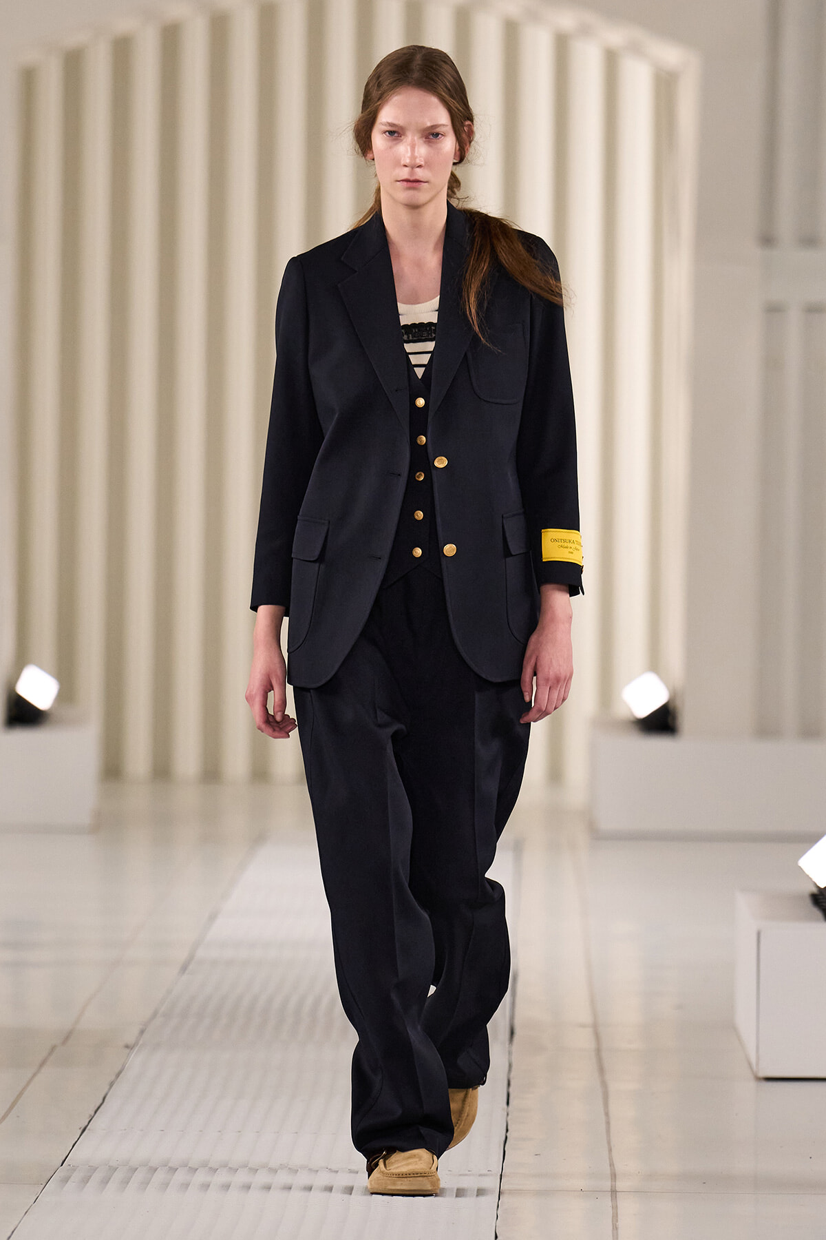 Female model walking a runway in a navy suit with gold buttons and tan shoes