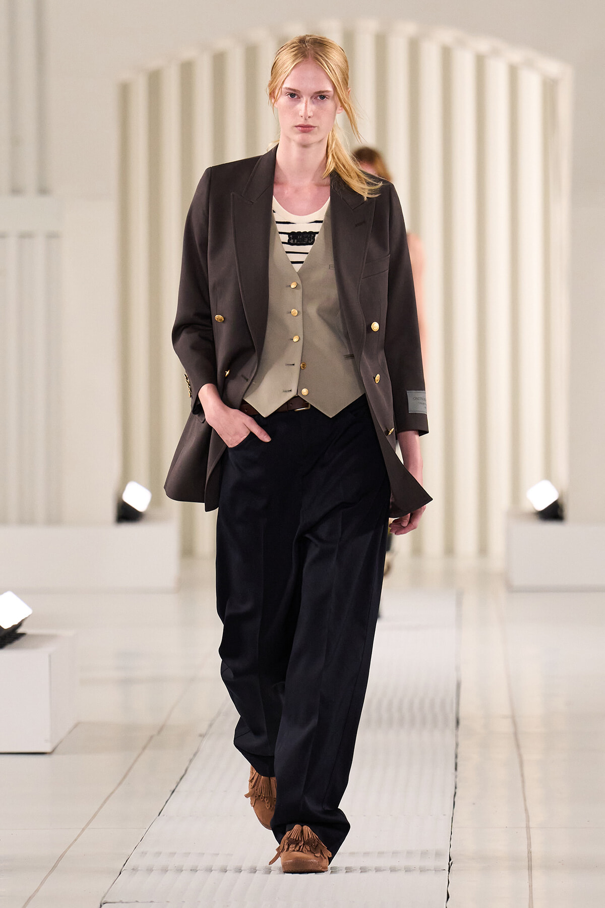 Female model walks a runway in a dark blazer over a tan waistcoat and striped top, with wide black trousers and tan fringe shoes.