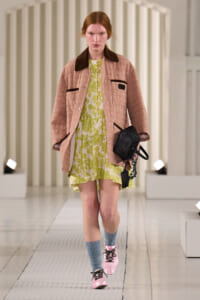 Model walking the runway in a pink quilted jacket over a lime floral dress, with pink lace-up shoes and blue socks, carrying a black studded bag.