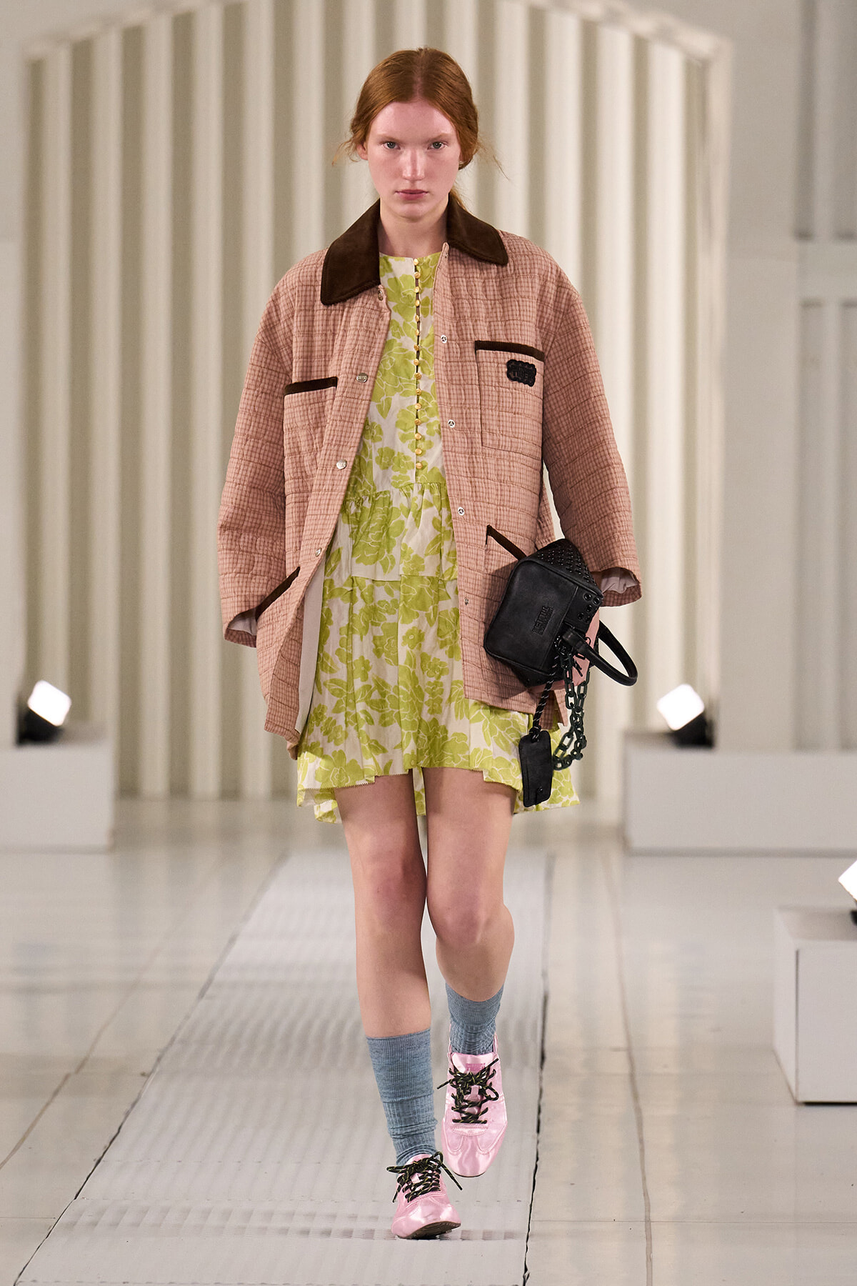 Model walking the runway in a pink quilted jacket over a lime floral dress, with pink lace-up shoes and blue socks, carrying a black studded bag.