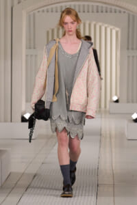 Young model walking runway in a layered pastel hoodie, gray scalloped dress, and coral-pink quilted jacket with cream accents
