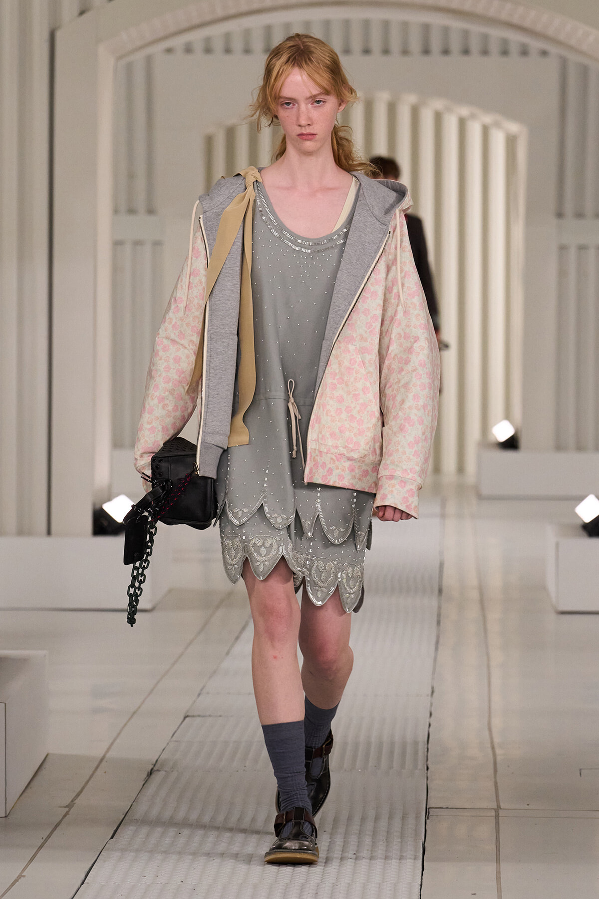 Young model walking runway in a layered pastel hoodie, gray scalloped dress, and coral-pink quilted jacket with cream accents