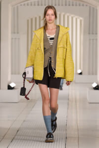 Runway model wearing a bright yellow textured coat over a gray vest, striped top, black shorts, blue socks, sandals, and a white handbag with a red chain.
