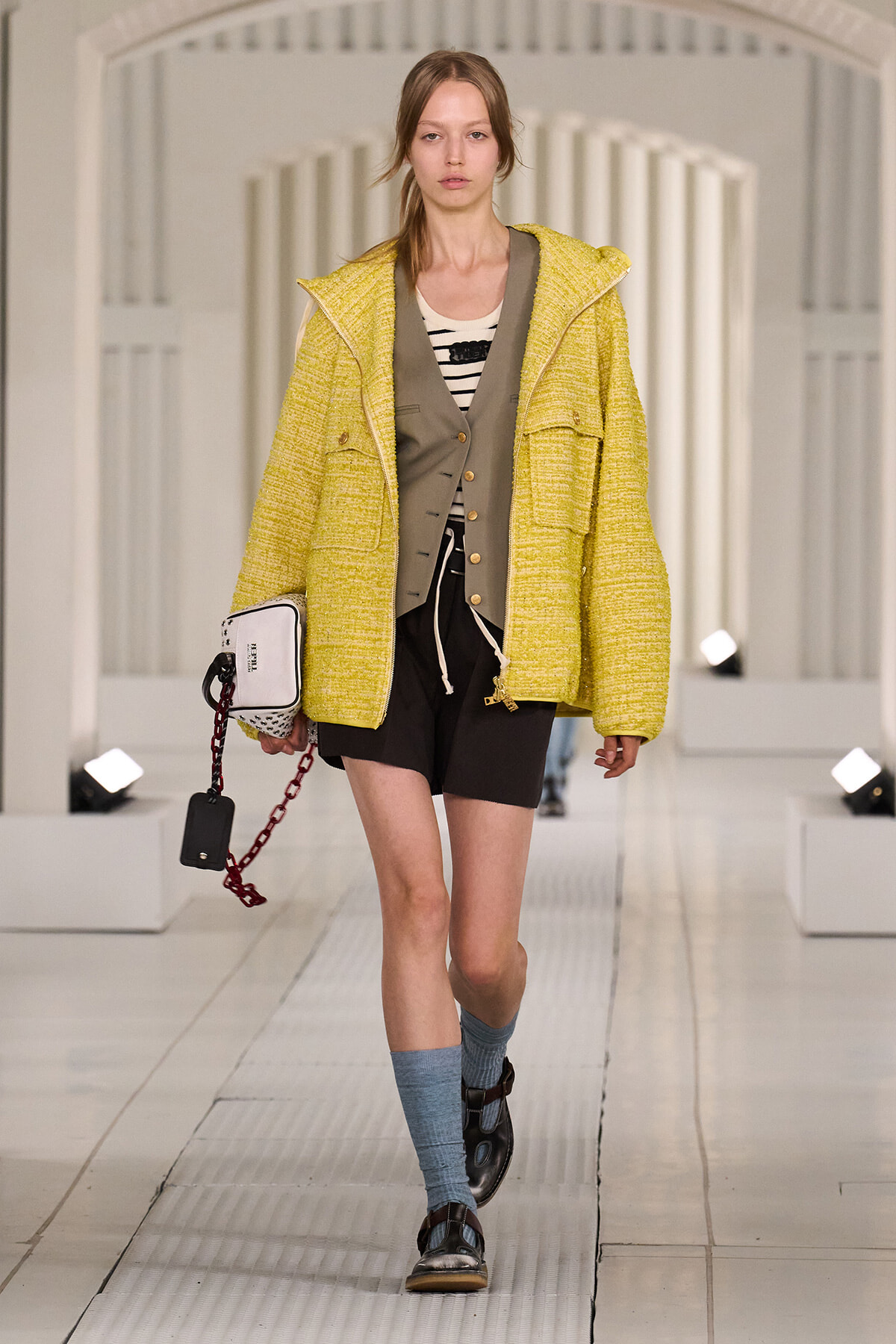 Runway model wearing a bright yellow textured coat over a gray vest, striped top, black shorts, blue socks, sandals, and a white handbag with a red chain.