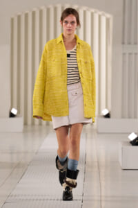 Model walks a runway in a bright yellow textured jacket over a striped top and white mini skirt, with blue socks and black boots.