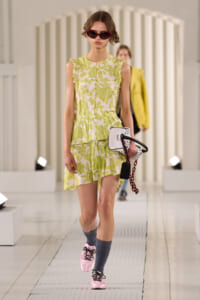 Model walking a runway in a lime green floral sleeveless dress with ruffled edges, pink sunglasses, and pink sneakers, carrying a white handbag with red chain accessory.