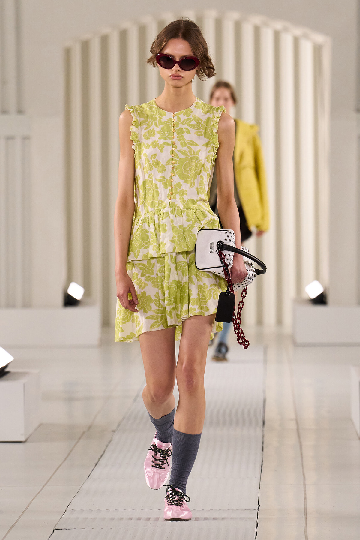 Model walking a runway in a lime green floral sleeveless dress with ruffled edges, pink sunglasses, and pink sneakers, carrying a white handbag with red chain accessory.