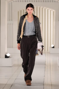 Model walking a runway in a gray ruffled top, textured black jacket, dark wide-leg pants, and brown fringe shoes, carrying a white handbag with a chain.