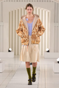 Model walking a white runway in a beige floral hooded jacket over a lavender blouse and beige pleated skirt, paired with olive socks and dark buckled loafers.