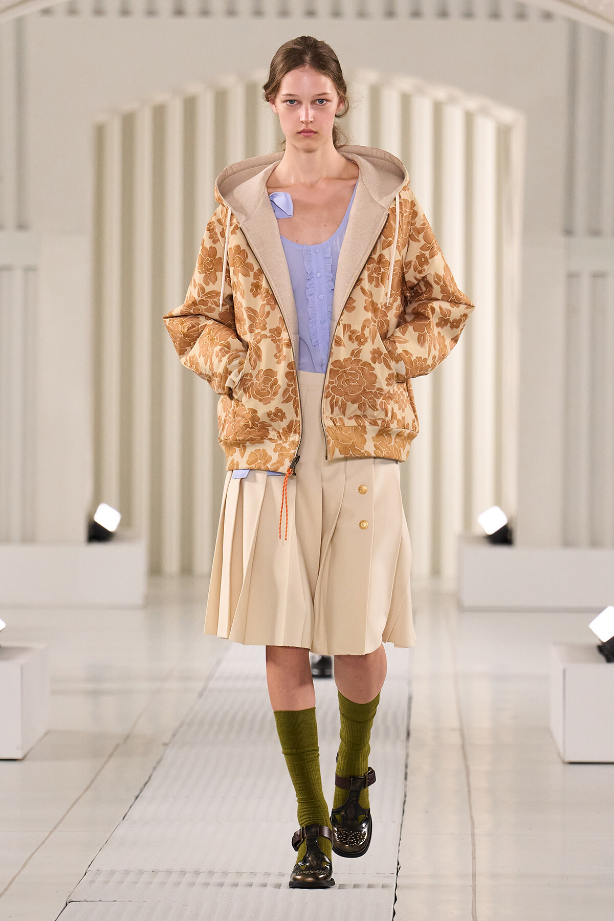 Model walking a white runway in a beige floral hooded jacket over a lavender blouse and beige pleated skirt, paired with olive socks and dark buckled loafers.