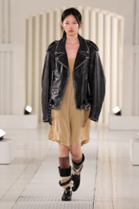 Model on a runway wearing a black oversized leather jacket over a beige lace-trim slip dress, with layered boots and socks.