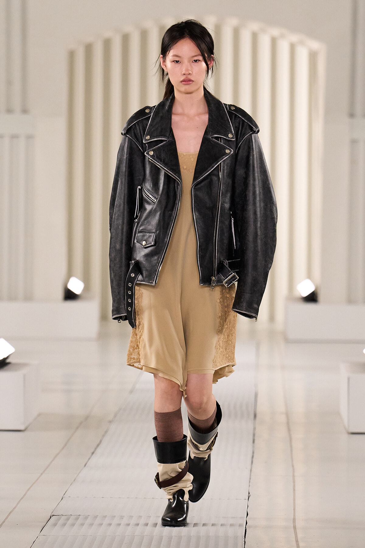 Model on a runway wearing a black oversized leather jacket over a beige lace-trim slip dress, with layered boots and socks.