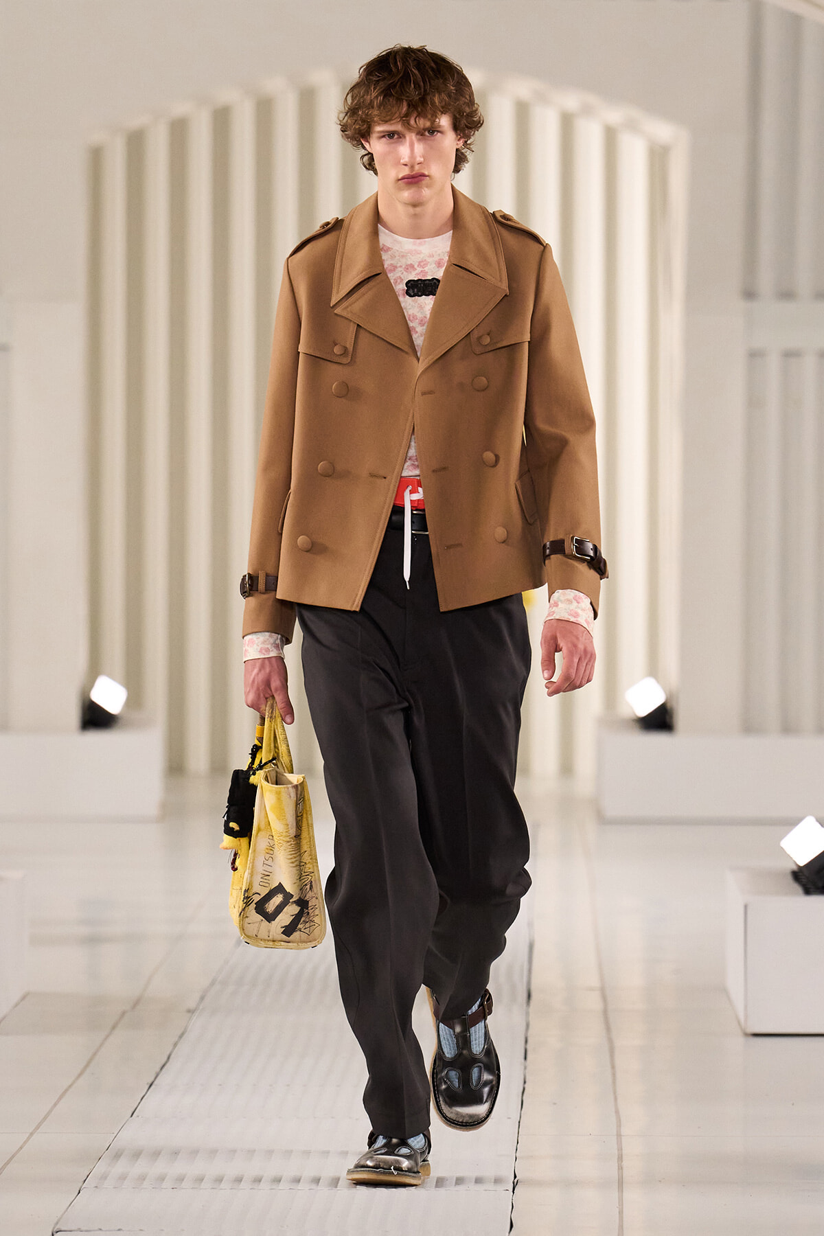Male model walking a runway in a tan double-breasted coat, pink patterned shirt, dark trousers, and sandals, carrying a yellow tote bag.