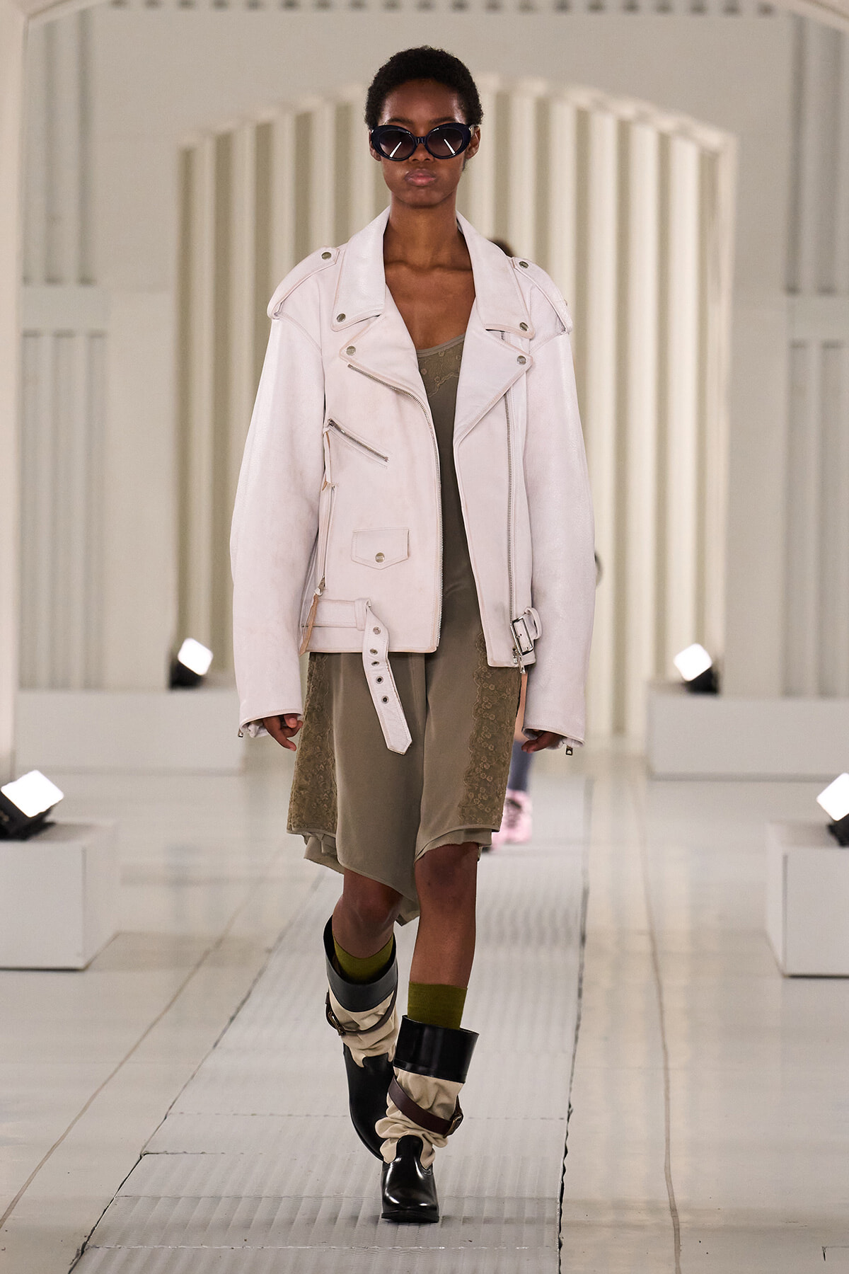 Model on a runway wearing a pale pink oversized biker jacket over an olive dress, with dark sunglasses on.