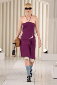 Model walking a fashion show in a strapless purple dress with a white crescent accent, beige underlayer, blue socks, and black sandals, carrying a brown bag with chain accessory and a phone.